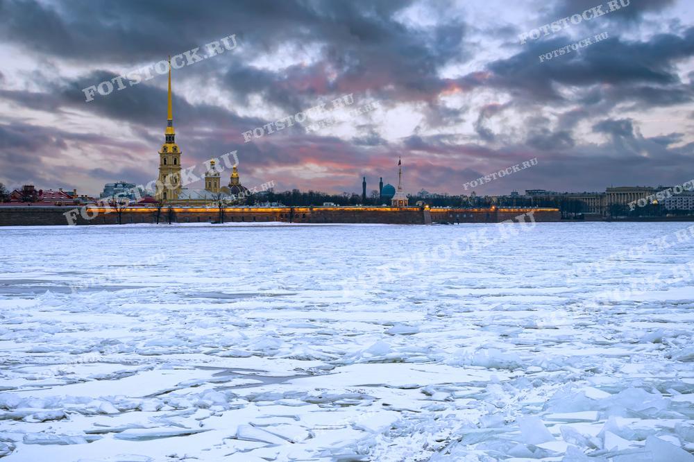 На фотографии изображена Петропавловская крепость в зимний вечер. Ледоход на реке Нева подчеркивает холодный сезон в Санкт-Петербурге. Вечернее небо с облаками добавляет драматичности этому историческому месту. Петропавловская крепость освещена огнями, создавая впечатляющий контраст с замерзшей рекой.